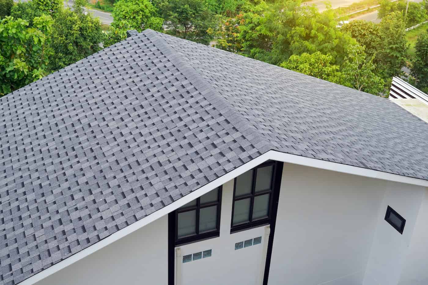 High-angle view of a modern house's steep gray asphalt shingle roof, surrounded by lush green trees.