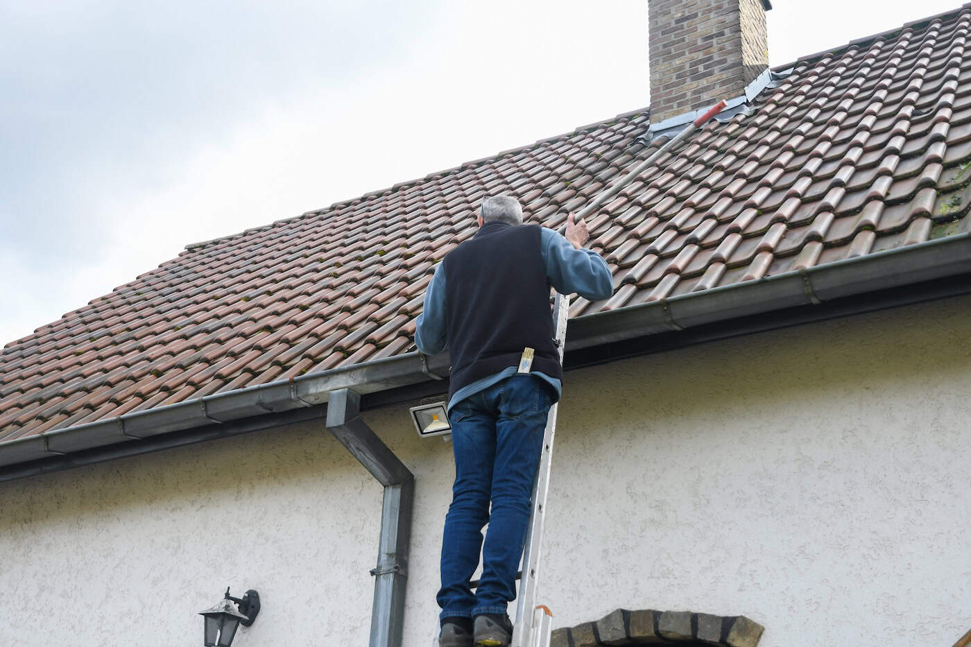 Roofer cleans gutter with cleaning tool while standing on ladder, performing necessary maintenance to prevent roof leaks and ensure proper drainage of rainwater